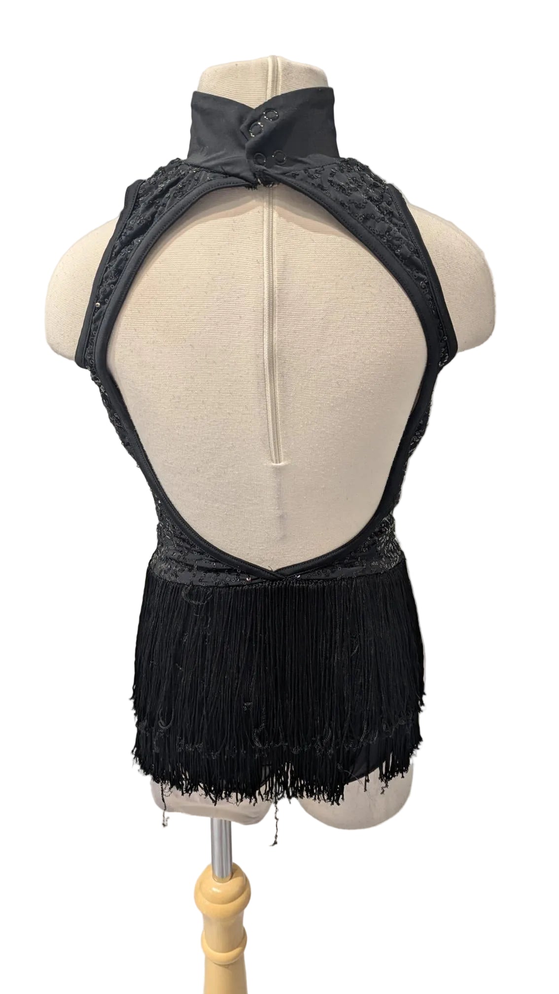 A black halter-neck dance dress with a high-cut fringe hem, displayed from the back on a cream-colored dress form mannequin against a white paneled wall.