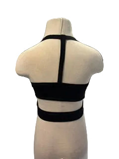 A black strappy crop top displayed on a beige mannequin torso, showing the back view with three horizontal straps and a central vertical strap. The setting appears to be a boutique interior with a gray floor, blue curtains, and a mirror in the background.