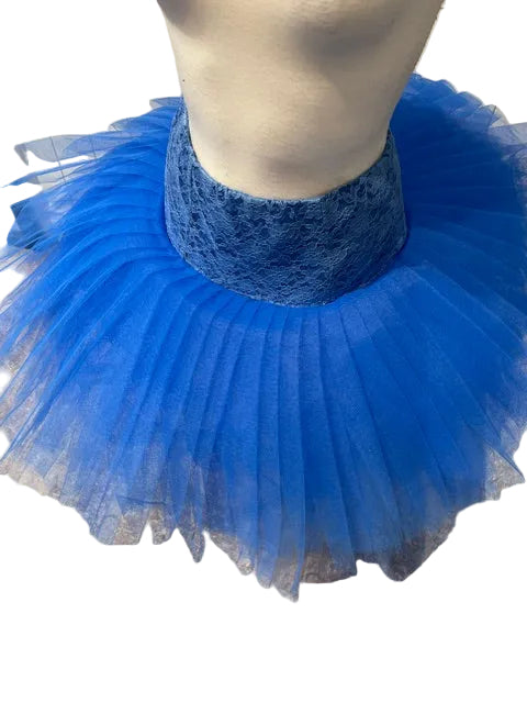 A bright blue tutu skirt displayed on a mannequin torso, featuring a wide blue lace waistband and layered tulle flares, set against a light wood-paneled wall and gray floor.