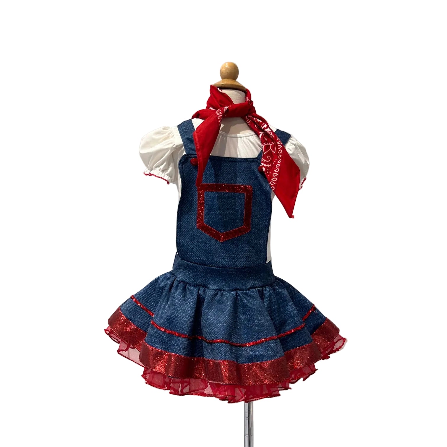 A children's denim overall dress displayed on a mannequin against a white paneled wall. The dress has a red pocket trim, a red ruffled hem, a white short-sleeve shirt underneath, and a red bandana tied around the neck.