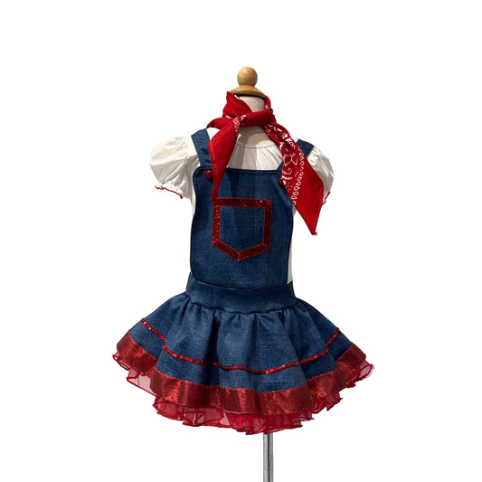 A children's denim overall dress displayed on a mannequin against a white paneled wall. The dress has a red pocket trim, a red ruffled hem, a white short-sleeve shirt underneath, and a red bandana tied around the neck.