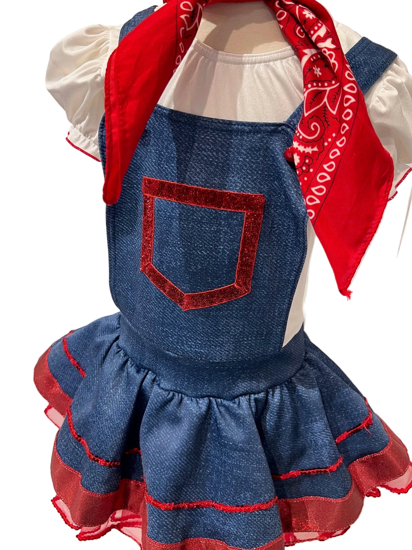 A children's costume dress displayed on a mannequin torso, featuring a blue denim-look overall-style bodice with a red glittery pocket detail, a white short-sleeve top underneath, and a red bandana tied around the neck. The skirt is blue with red layered trim.