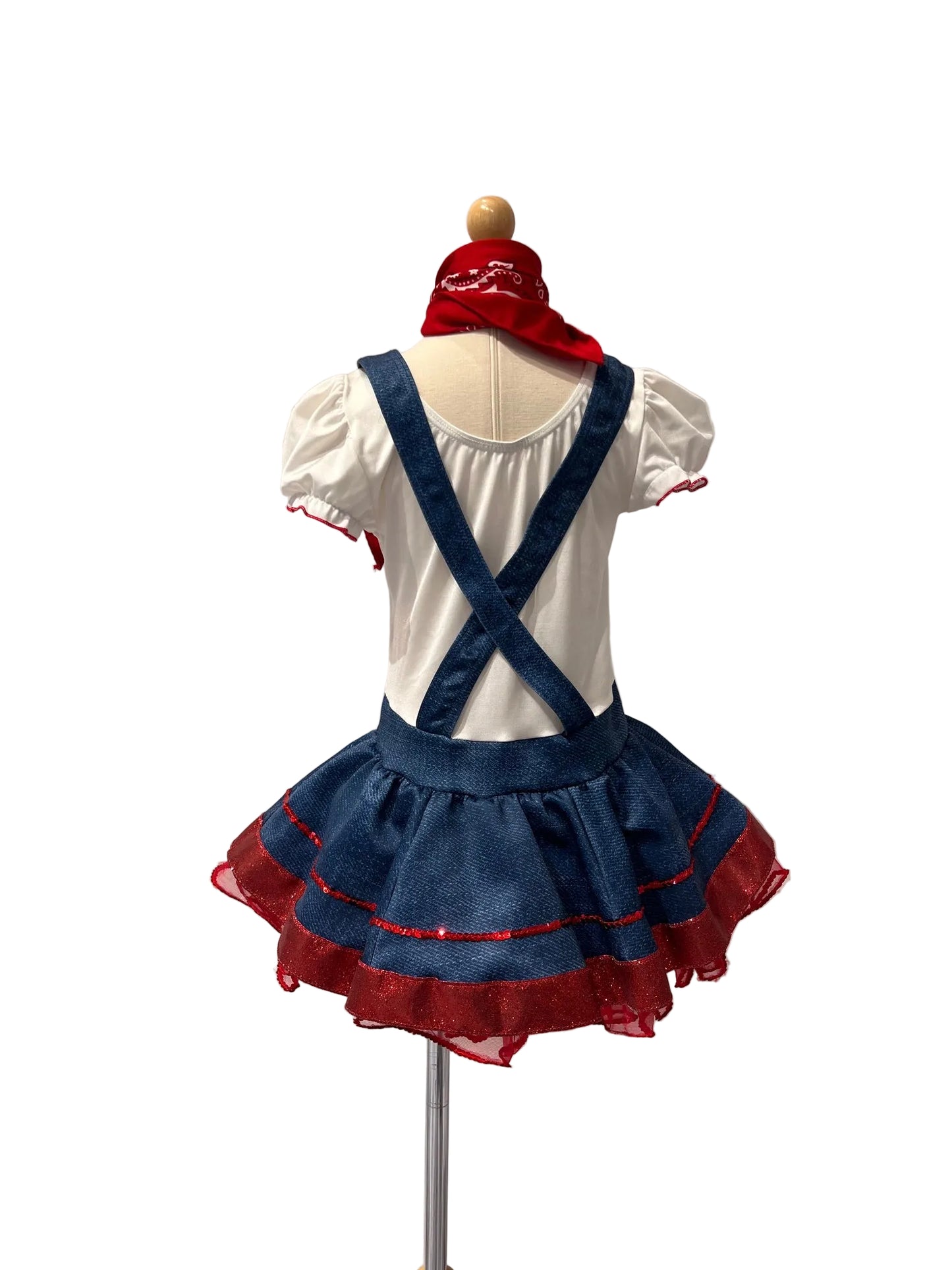 A children's costume displayed on a mannequin from the back, featuring a white short-sleeve top with red trim, blue denim-style suspenders, a blue ruffled skirt with red mesh trim, and a red headband.