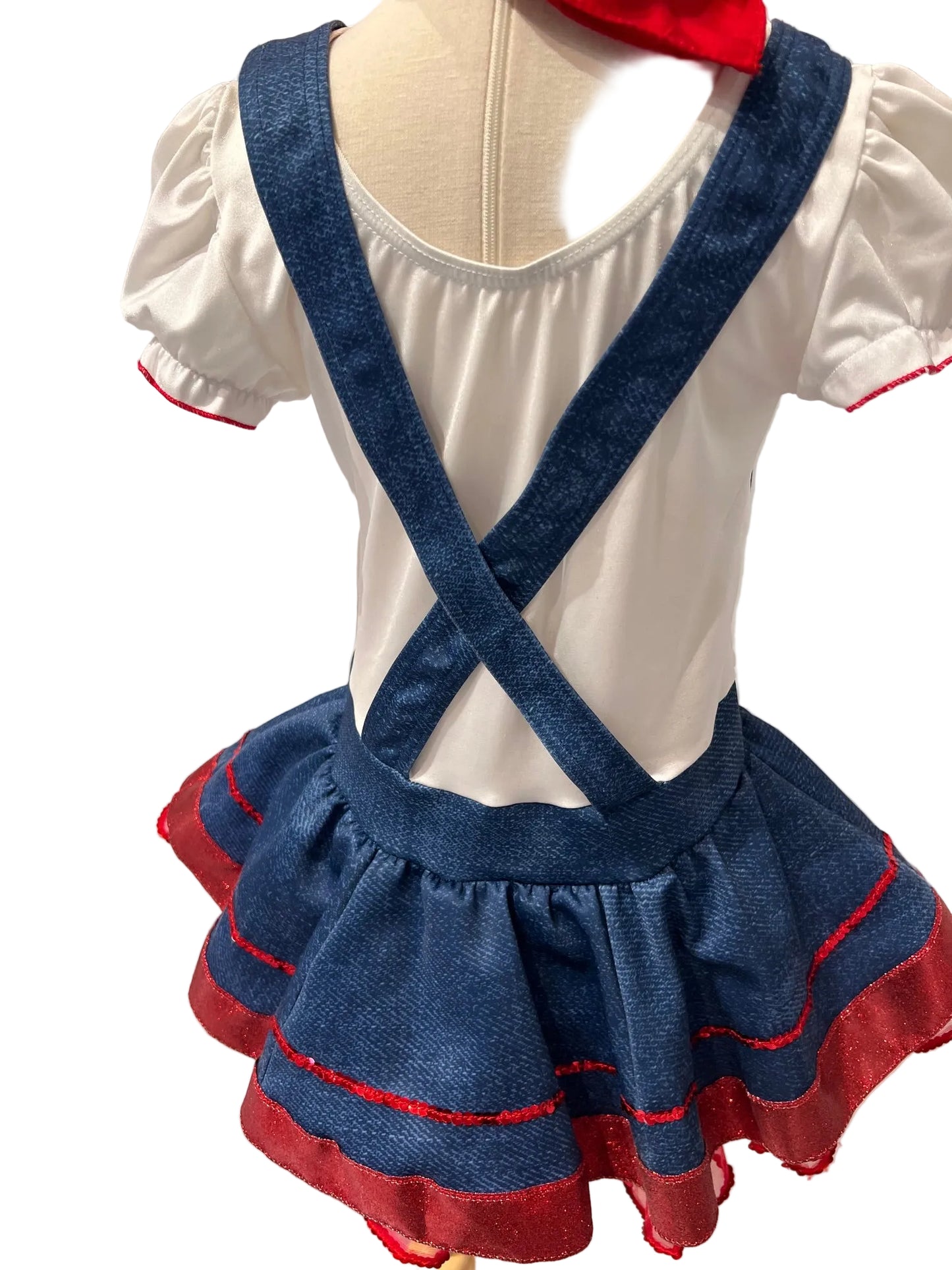 A children's dress displayed on a mannequin, featuring a white short-sleeve top with red trim, blue denim-style suspenders, and a ruffled blue skirt with red trim, set against a white paneled wall.