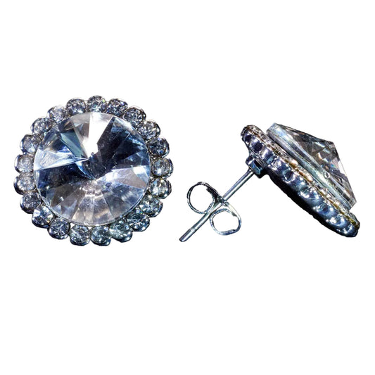 A pair of round halo stud earrings featuring large clear faceted center stones surrounded by a border of smaller clear stones, shown on a dark textured background. One earring is facing forward, and the other is angled to show the post and backing.