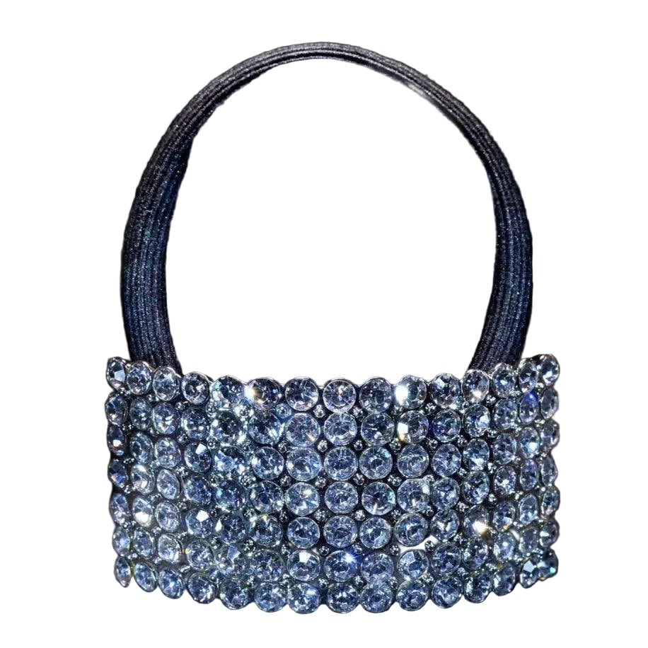 A small structured handbag with a wide, heavily embellished top band of clear rhinestones and a single black elastic handle, photographed against a dark studio background.