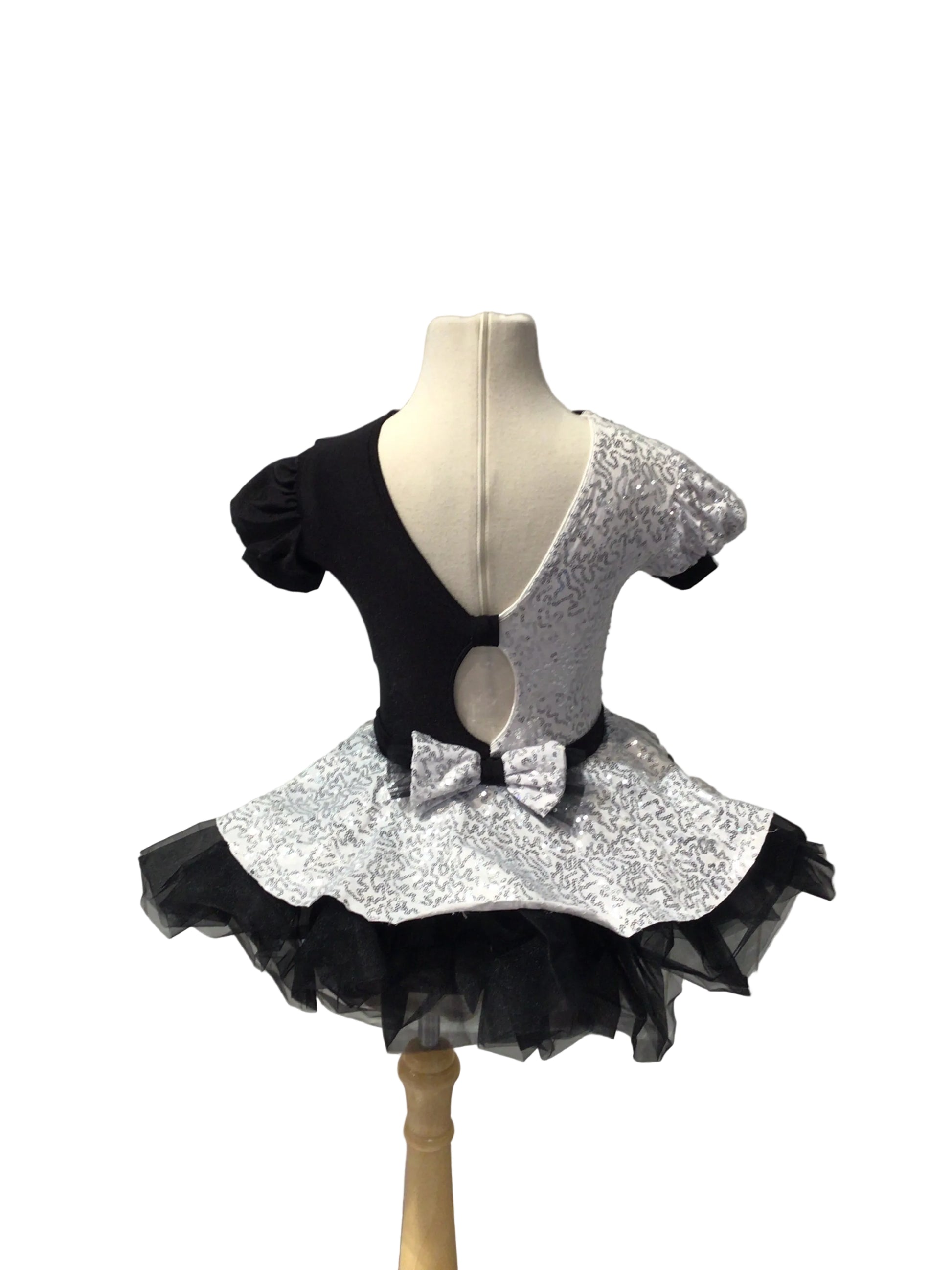 A black and silver sequined dance dress displayed on a mannequin torso, featuring a split black and silver bodice, short puff sleeves, a bow at the waist, and a layered black tulle tutu skirt, set against a white paneled wall.