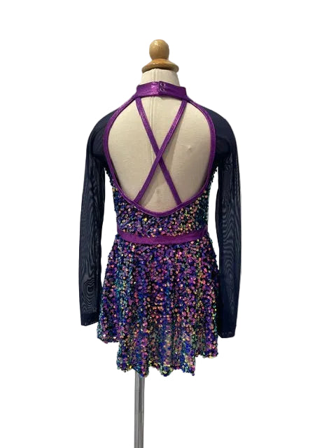 A girls' figure skating dress displayed on a mannequin from the back, featuring a purple halter-style back with crisscross straps, a purple waistband, and a skirt covered in multicolored sequins, with dark sheer long sleeves.